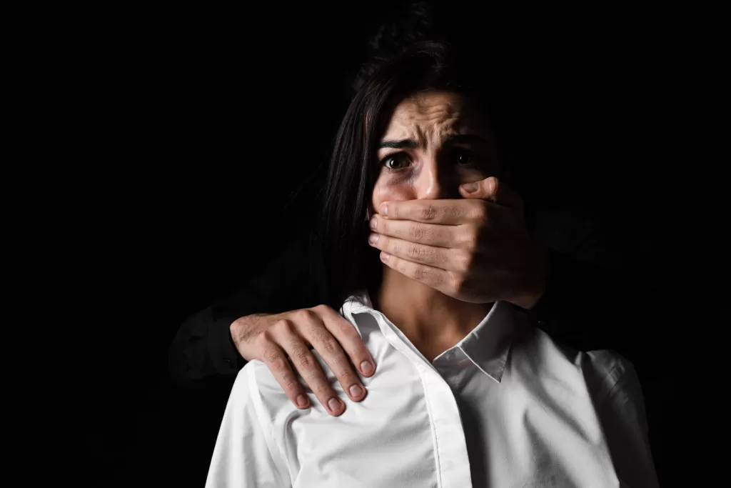 A man standing behind a woman in the dark with is hand sitting menacingly on her shoulder and a hand over her mouth. If you are experiencing domestic violence you need to contact a Kansas City domestic violence lawyer to help take control back of your life.